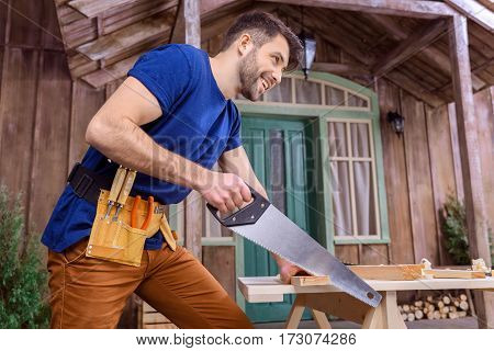 Low angle view of smiling bearded carpenter in tool belt sawing wooden plank on porch