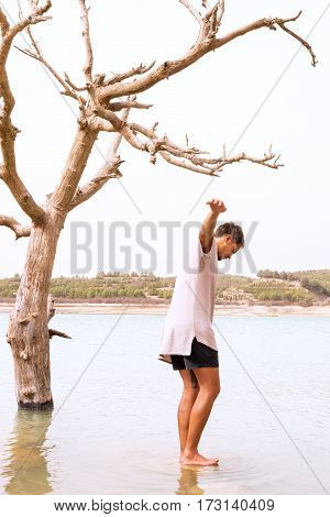 young male balancing on a rock in a flooded lake with a dead tree in the water. concept of climate change filter added blank sky in background for copy space