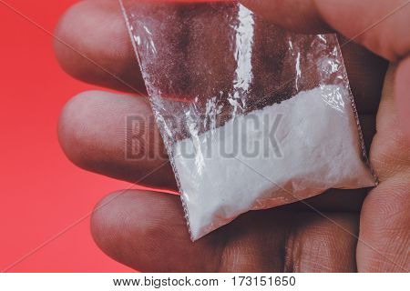 Mans hand holding or giving, selling on palm plastic packet with cocaine powder or another drugs on red background. Close-up macro retro toned photo.