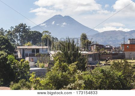 View of volcano from rural Guatemalan town. Partly cloudy blue sky above with houses green bushes and trees.