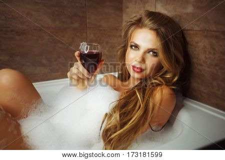 Young cute girl drinking red wine in bath with foam. She relaxes after a hard day. She coyly looking at the camera.