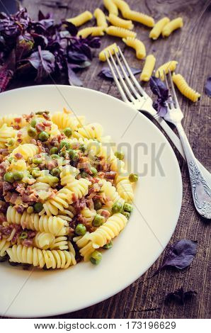 Fusilli with prosciutto and green pease on old wooden background. Traditional Italian pasta with fresh basil. Selective focus.