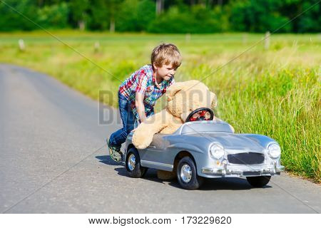 Little preschool kid boy driving big toy car and having fun with playing with his plush toy bear, outdoors. Child enjoying warm summer day in nature landscape