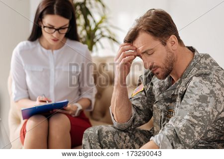 Coping with stress. Cheerless handsome bearded man holding his forehead and closing his eyes while trying to cope with posttraumatic stress