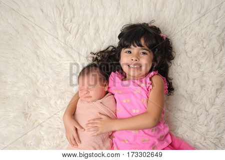 Portrait of a 3 1/2 year old sister holding her 9 day old baby sister. Shot from overhead on a white flokati rug.