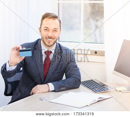 Toned of happy toothy smiling businessman holding credit card while sitting in front of computer. Businessman in black suit offering advantageous agreement or contract.