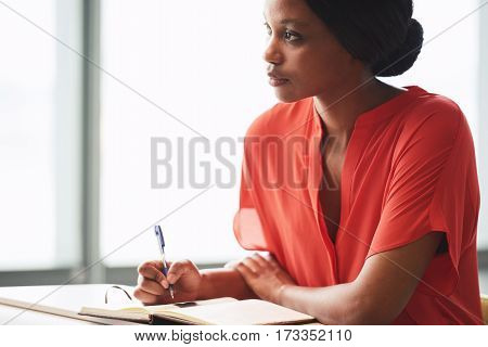Black female creative writer busy looking up into the distance as she procrastinates and takes a break from her writing session while dressed in a colourful orange shirt.
