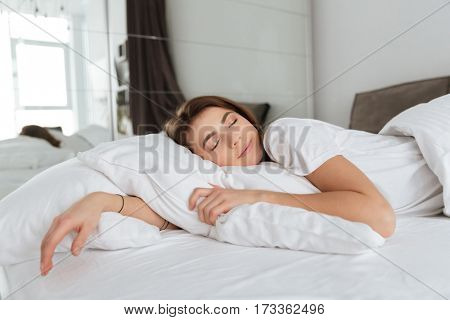 Picture of young tired smiling woman dressed in white t-shirt lies in bed at home indoors and sleeping.