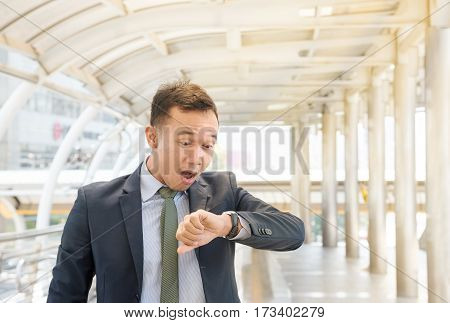 Portrait of worried young Asian businessman looking at his watch checking time for meeting in shock and horror with wide eyes mouth agape as he stares at wristwatch copy space.