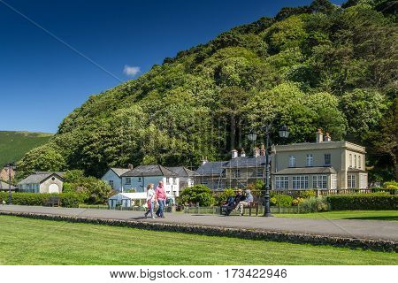 Lynmouth Devon England 13 July 2016: People walk along the path past the small houses otlele and cafes. Two sitting on the bench. Sea shore.