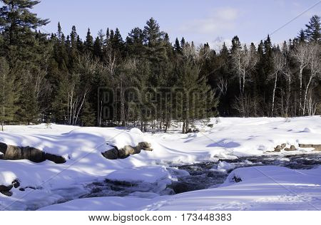 Wide view of a snowy country landscape with a small stream of rapids running through and a background of green fir trees at Pabineau Falls near Bathurst, New Brunswick, on a bright sunny day with blue skies and clouds in February.