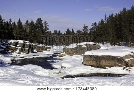 Wide view of a snowy country landscape with a small stream of rapids running through rock faces on either side and a background of green fir trees at Pabineau Falls near Bathurst New Brunswick on a bright sunny day with blue skies and clouds in February.