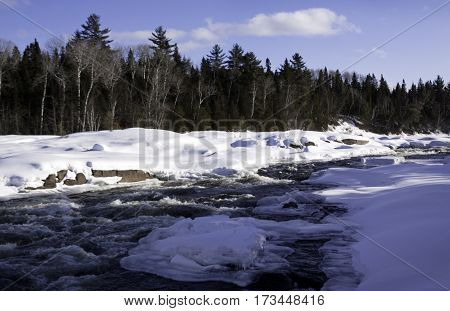Wide view of a snowy country landscape with a small stream of rapids running through and a background of green fir trees at Pabineau Falls near Bathurst, New Brunswick on a bright sunny day with blue skies and clouds in February.