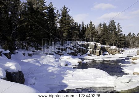 Wide view of a snowy country landscape with a small stream of rapids running through a large rock face on the side and a background of green fir trees at Pabineau Falls near Bathurst New Brunswick on a bright sunny day with blue skies and clouds in Februa