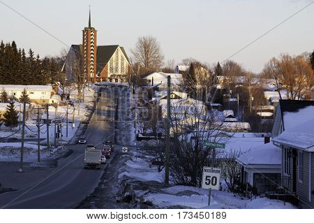 Wide view of a long street with vehicles leading up a hill to a church at the top with a houses and buildings on either side in Bathurst New Brunswick on a bright sunny day with blue skies and clouds in February.