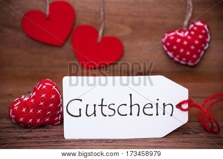 Label With German Text Gutschein Means Voucher. White Label With Red Textile Hearts. Retro Brown Wooden Background.