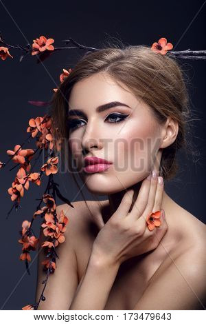 Beautiful young woman with make-up and loose hairdo holding artificial sakura branch with orange flowers. Beauty shot on dark background. Copy space.