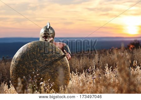 Ancient Greek soldier holding a sword hiding behind his shield on the battlefield ready to attack copyspace. Spartan warrior on the battlefield beautiful sunset on the background bravery hero concept