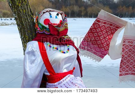 Shrovetide doll in colorful headscarf and sarafan near tree on background of spring landscape