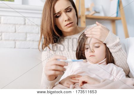 So worried. Troubled cheerless mother measuring temperature of her little sick daughter while sitting together on the sofa