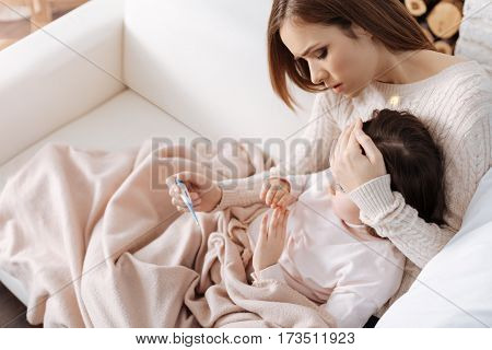 Urgent threat. Cheerless caring mother sitting on the sofa and measuring temperature of her sick daughter while helping to recover