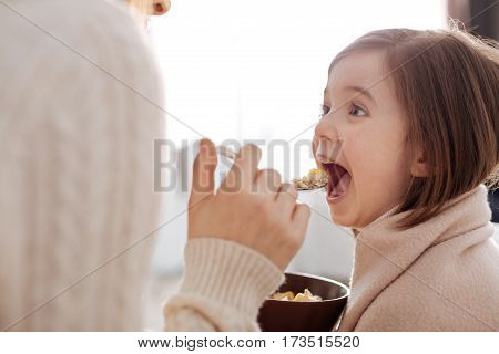 Yammy yammy. Pleasant caring mother feeding her daughter with breakfast while resting together on the sofa
