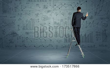 A businessman in modern stylish elegant suit standing on a small ladder and drawing pie and block charts on grey wall background with exponential progressing curves, lines, angles, blocks, numbers