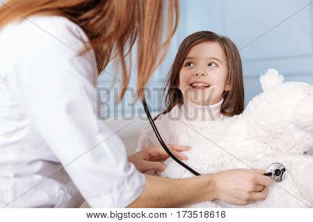 Everything is ok. Pleasant professioanl doctor examining fluffy bear with stethoscope while little smiling girl looking at her