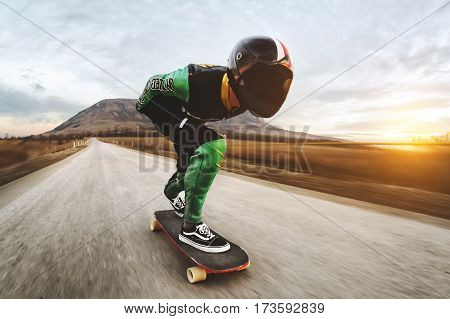 A man in a fullface helmet and a leather suit in a special rack rides a longboard on afsaltu sunset in the background mountains and beautiful sky at high speed.