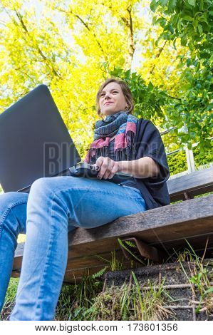 Young woman using laptop at a riverbank. Milleanial working in a park.