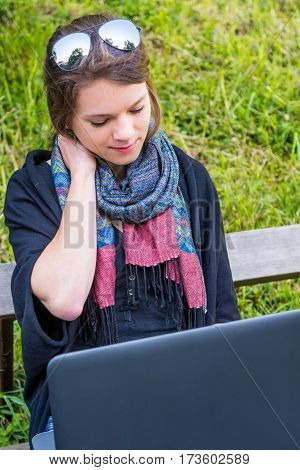 Young woman rubbing her neck while working on a laptop. Millenial working in a park.