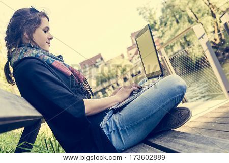 Young woman using laptop at a riverbank. Milleanial working in a park.