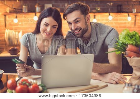 Order food online. Young beautiful couple in kitchen ordering food online from restaurant. Family of two using laptop. Young woman paying with credit card. Nice loft kitchen interior with light bulbs