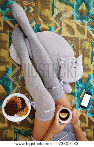 Girl sitting in bedroom in knee-height socks with a cup of tea waffels toy cat and a cell phone