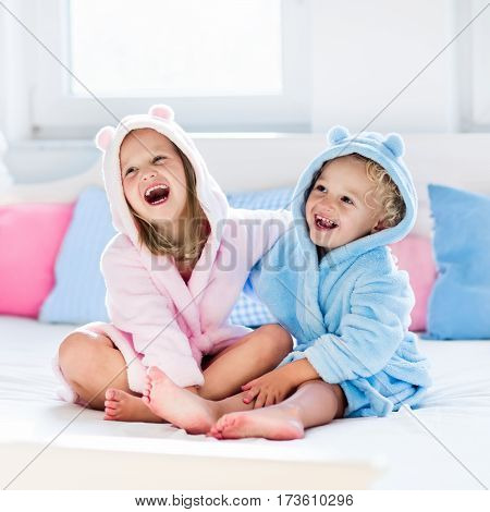 Happy laughing kids boy and girl in soft bathrobe after bath play on white bed with blue and pink pillows in sunny bedroom. Child in clean and dry towel. Wash infant hygiene health and skin care.