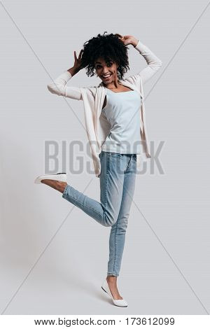 Playful woman. Full length of attractive young African woman in casual wear keeping hands in hair and looking at camera while standing against grey background