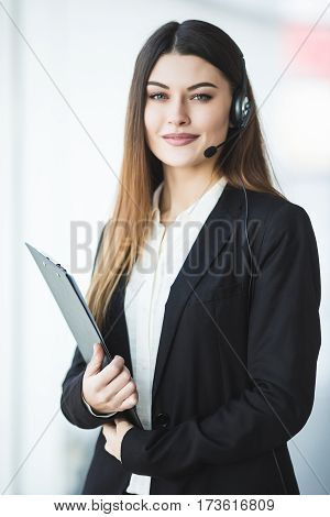 Photo of beautiful young call center operator standing in office. Woman with headphones looking at camera and smiling