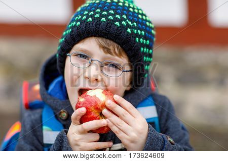 Little kid boy with eye glasses on the way to the school and eating fresh apple. Child on a street. Preschooler wearing warm clothes, glasses and backpack. Happy student or people. Education concept