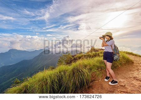 Hiker asian teen girl wear cap and eyeglass with happy holding dslr camera shooting photo beautiful nature in winter on peak mountain at sunset in Phu Chi Fa Forest Park Chiang Rai Thailand