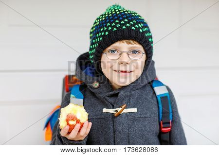 Little kid boy with eye glasses on the way to the school and eating fresh apple. Child with missing front teeth wearing warm clothes, glasses and backpack. Happy student or people. Education concept