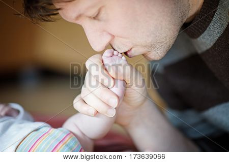 Father kissing foot of newborn baby. Young dad cuddling with new born child at home. Happy parenthood, carefree childhood, family, love, tenderness.