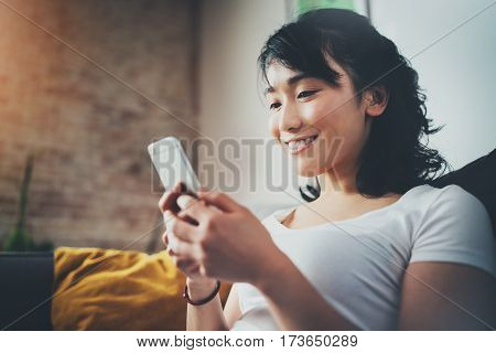 Young smiling Asian woman spending rest time at home and using smartphone for texting with friends.Selective focus. Blurred background, flares effect