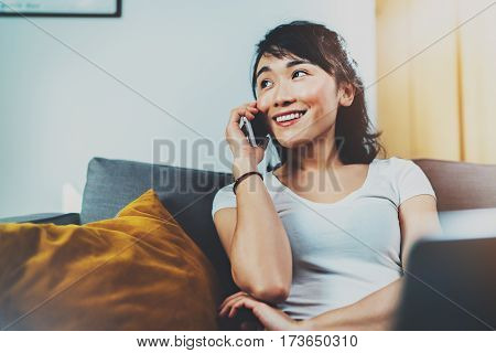 Young smiling Asian woman spending rest time at home on sofa and using smartphone for making call.Blurred background, flares effect