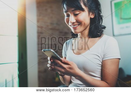 Happy young Asian woman holding mobile phone on hands and reading message at home room interior on the background. Flarres