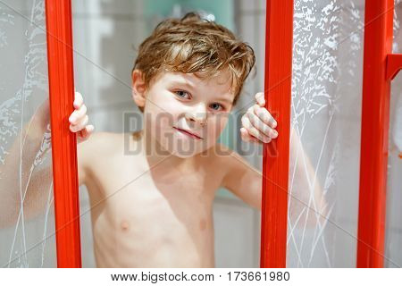 Happy little kid boy in the shower cabin wet and smiling. Adorable child with blond hairs taking bath and having fun.