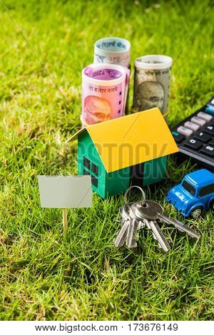 indian currency notes of rupees 100, 500 and 2000 in roll or stack with small home model, keys, toy car and calculator with blank board over green grass - home buying and finance concept in India