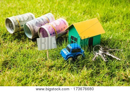 indian currency notes of rupees 100, 500 and 2000 in roll or stack with small home model, keys, toy car and calculator with blank board over green grass - home buying and finance concept in India
