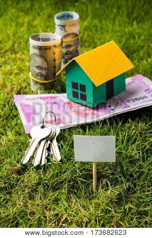 indian currency notes of rupees 100, 500 and 2000 in roll or stack with small home model, keys, toy car and calculator with blank board over green grass - home buying and finance concept in India