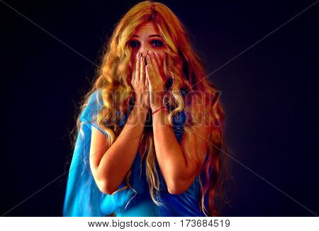 Frightened woman screaming with fear indoor at halloween night. Horror. Surprise and worried girl with tousled hair worried bawl and l covers her mouth with her hands black background.