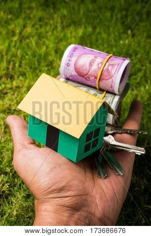 hand holding new indian currency notes or rupees 500 and 2000 and house model over green grass - indian real estate and finance concept